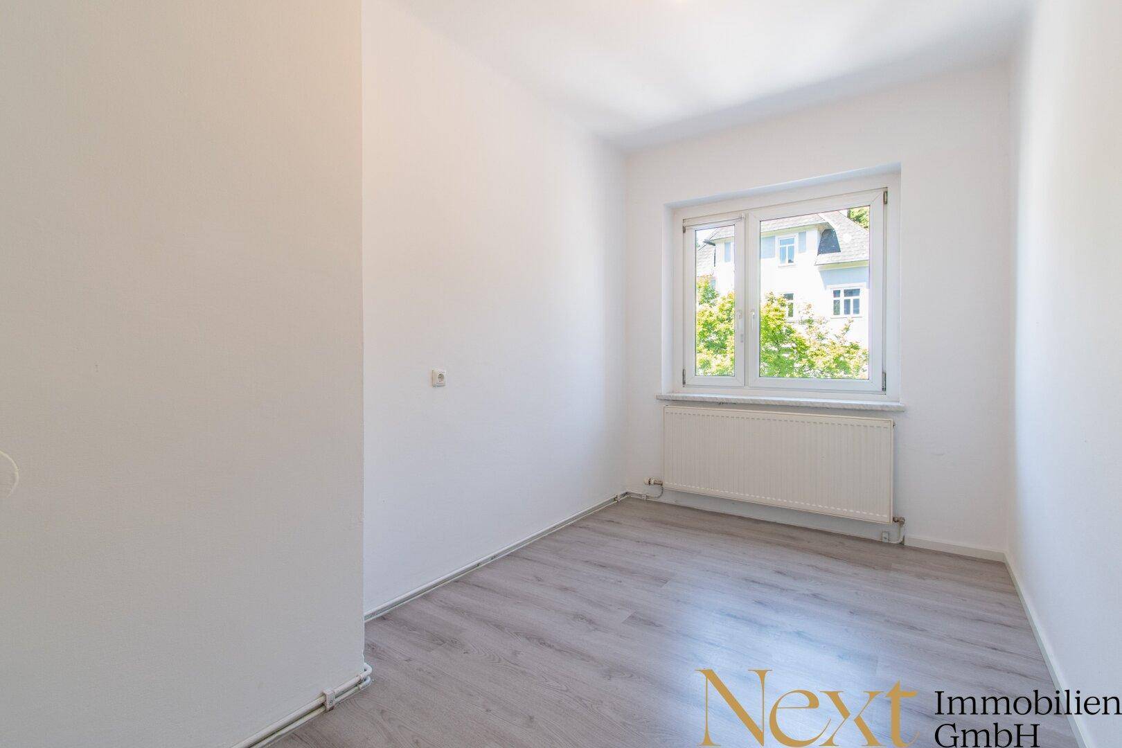 Gemütliches Schlafzimmer mit hellem Laminatboden, weißen Wänden und einem Fenster mit Blick auf Bäume.