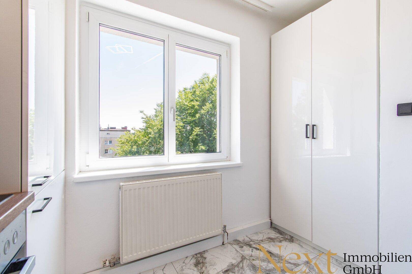Heller Küchenbereich mit Fenster, weißen Hochglanzschränken und Blick ins Grüne.