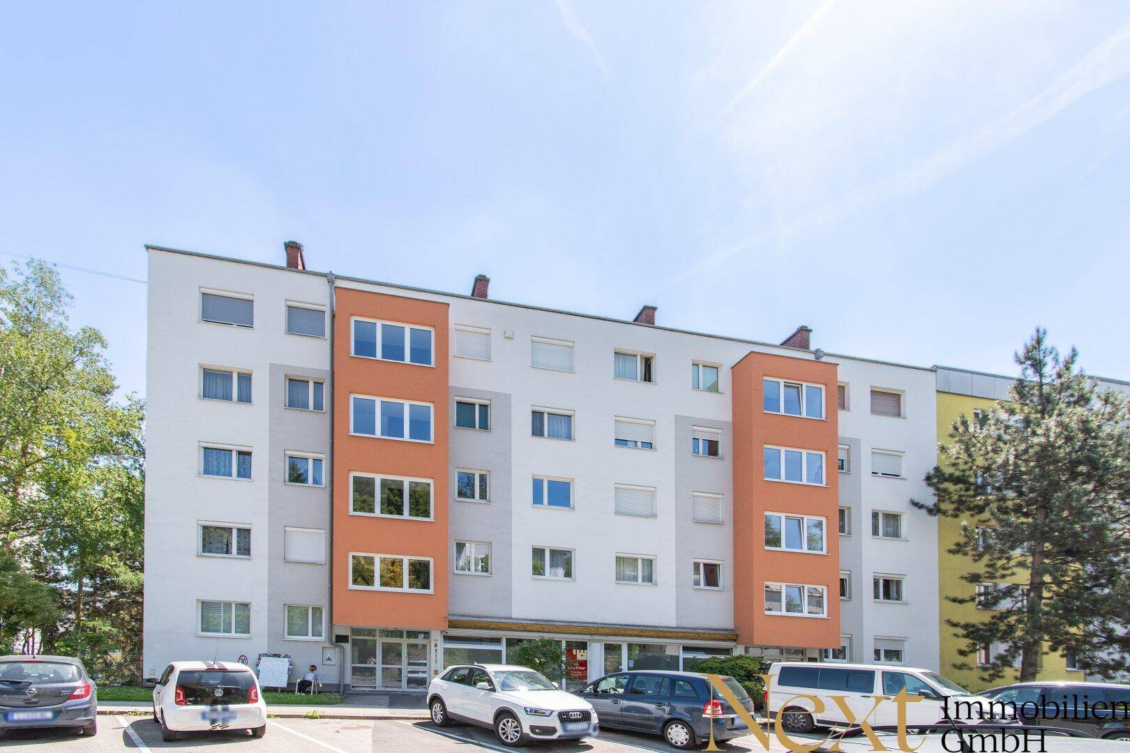 Mehrfamilienhaus mit moderner Fassade, Flachdach und Parkplätzen vor dem Gebäude.