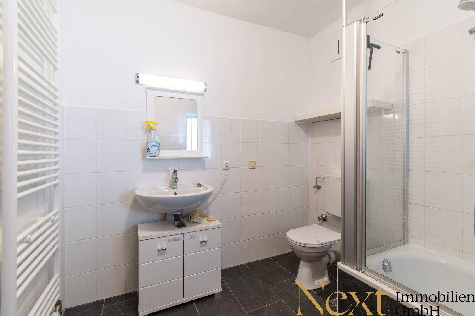 Badezimmer mit Dusche über Badewanne, Waschbecken und WC, gefliest in Weiß und Grau.