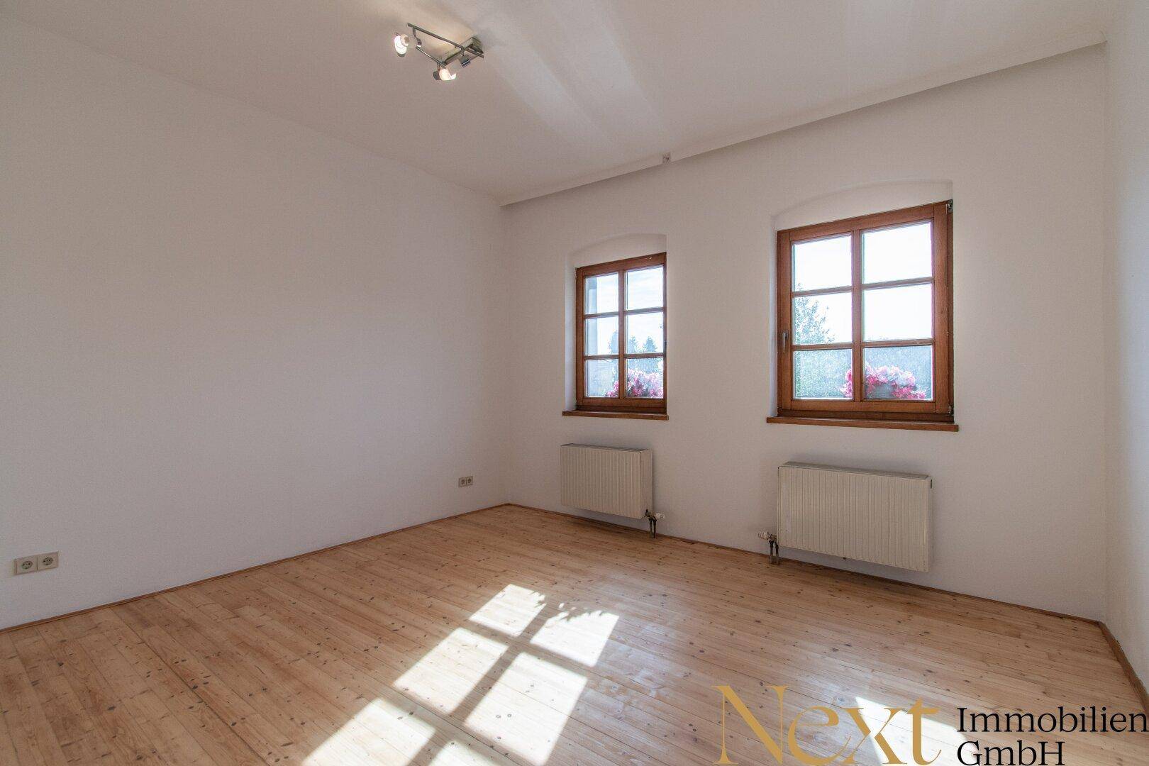 Helles Zimmer mit Holzdielenboden, zwei Fenstern und Heizkörpern, sonnendurchflutet.