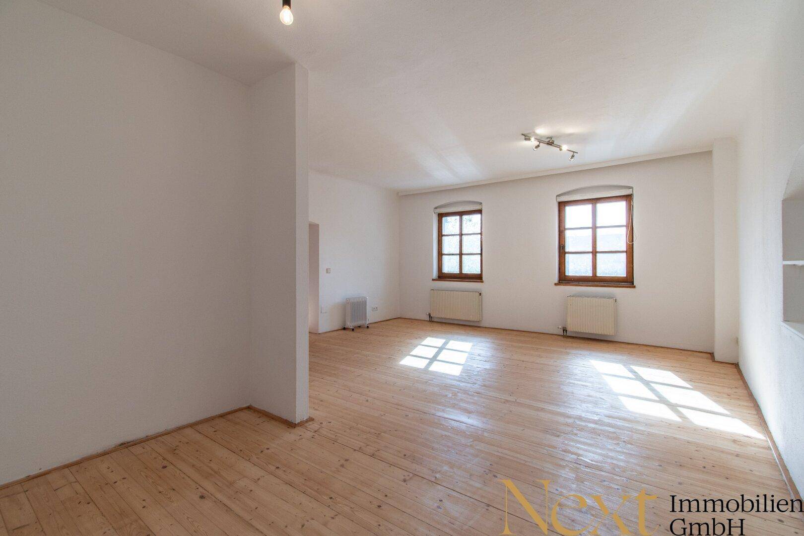 Großes, helles Zimmer mit Holzdielenboden und zwei Fenstern, die viel Tageslicht hereinlassen.