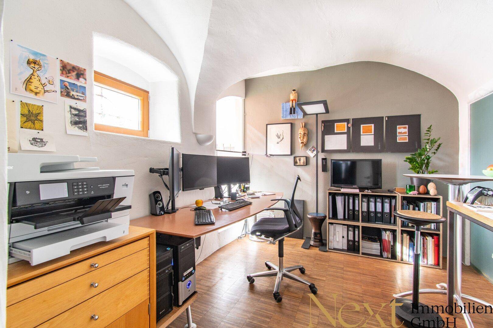 Geräumiges Heimbüro mit gewölbter Decke, Holzboden und zwei großen Monitoren am Schreibtisch.