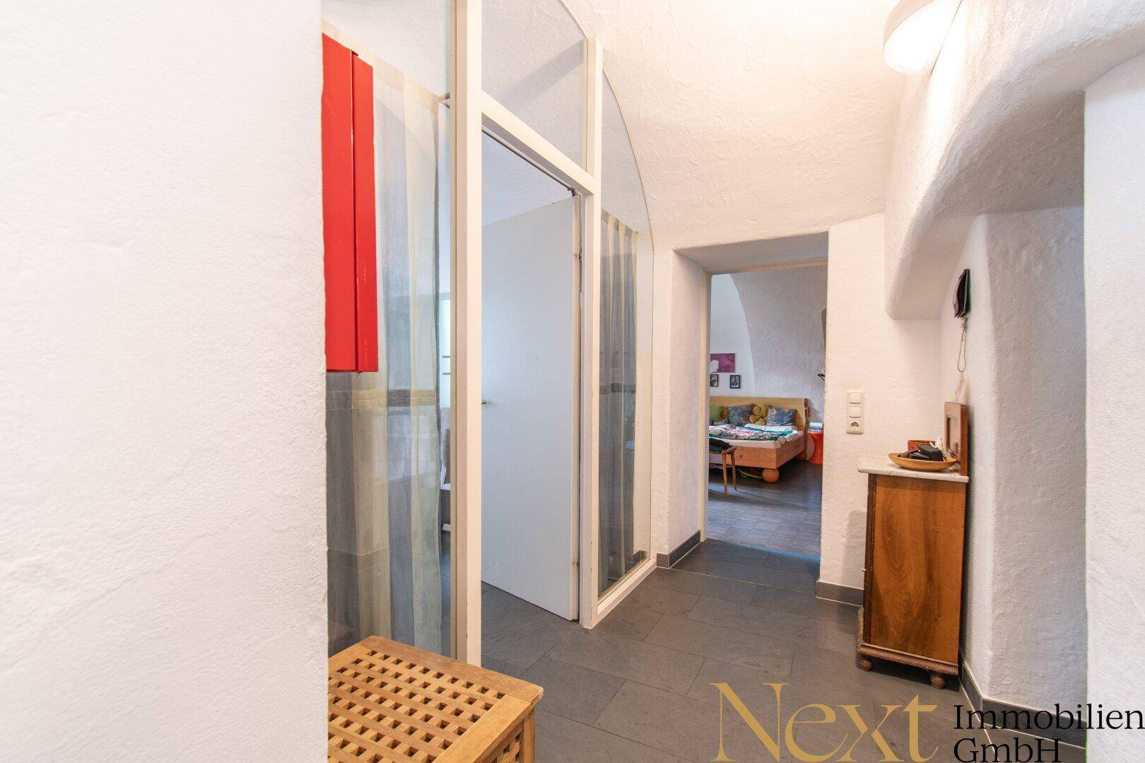 Heller Flur mit dunklen Fliesen, weiß verputzten Wänden und Durchgang zu einem Schlafzimmer.