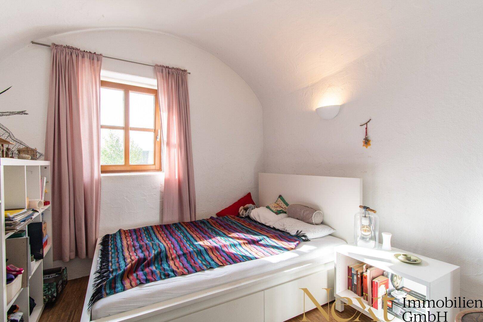 Gemütliches Schlafzimmer mit Holzbett, gewölbter Decke und Fenster mit rosa Vorhängen.