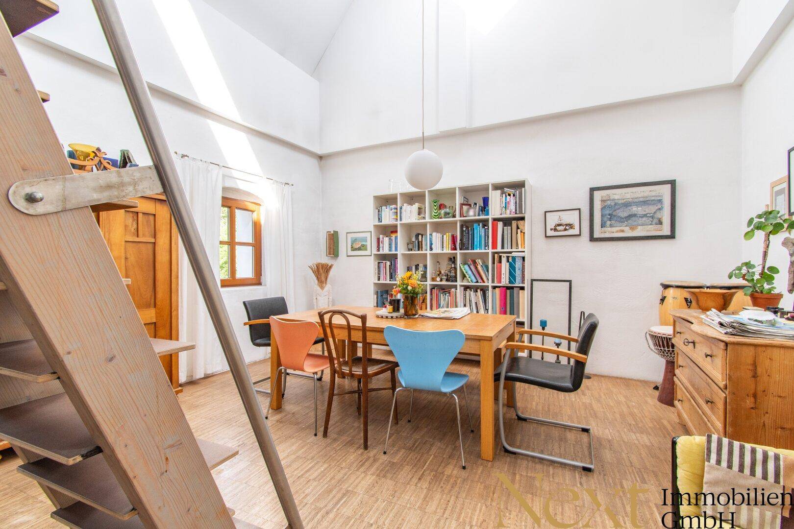 Heller Essbereich mit hoher Decke, Holztisch und Bücherregal, verbunden mit einer Galerie.