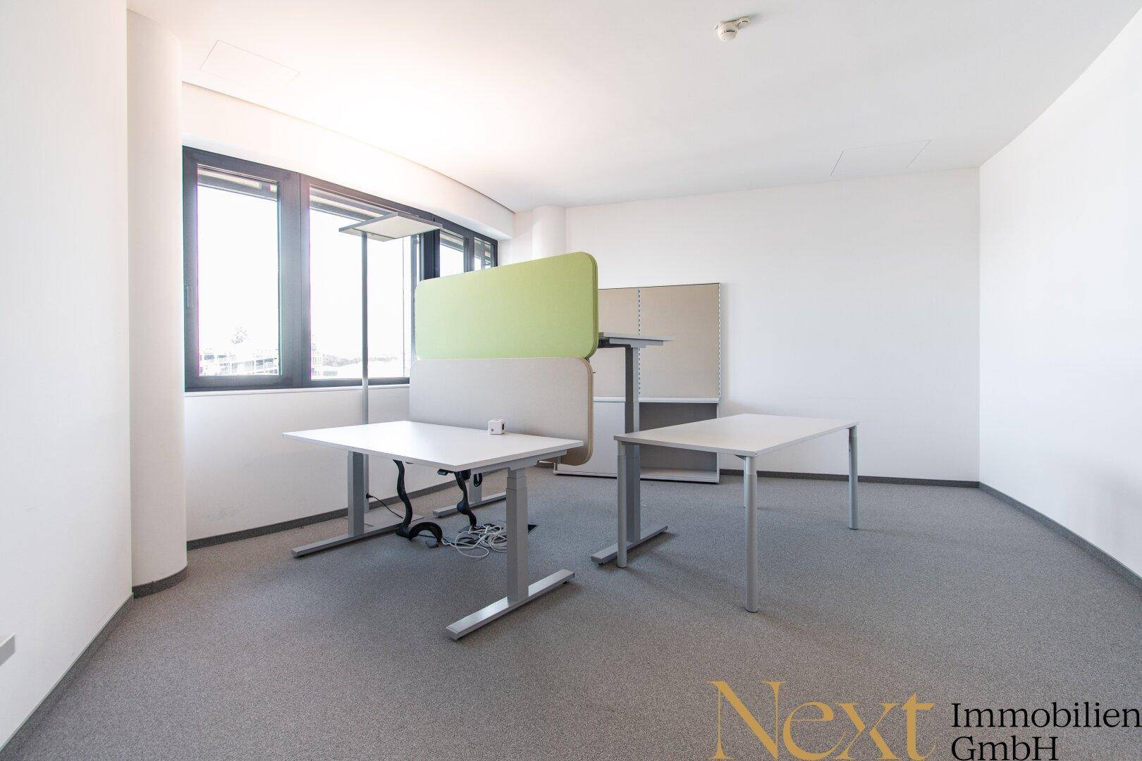 Leeres Büro mit zwei Schreibtischen, einem grünen Trennelement und großen Fenstern.