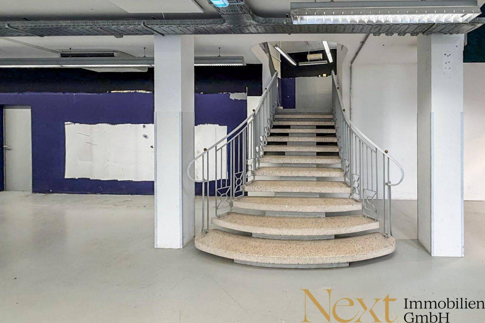 Geschwungene Treppe mit Metallgeländer in einem großen, offenen Gewerberaum mit Betonboden.