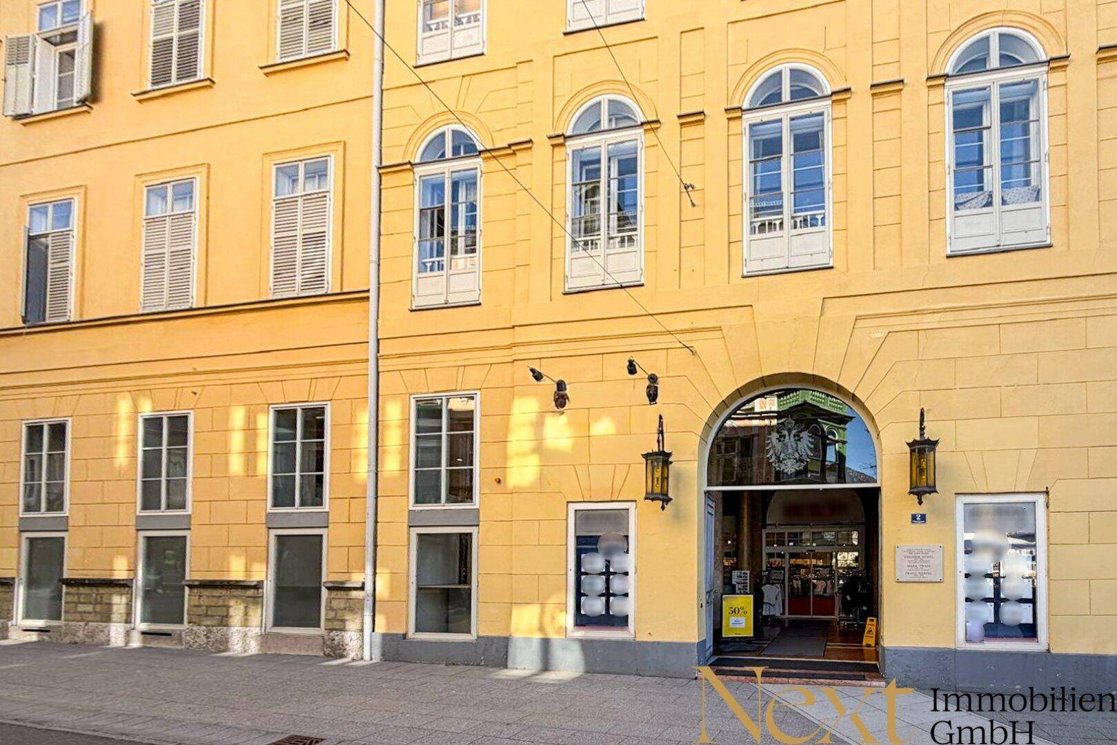 Gelbe Gebäudefassade mit großen Fenstern und einem markanten Torbogen als Eingangsbereich.