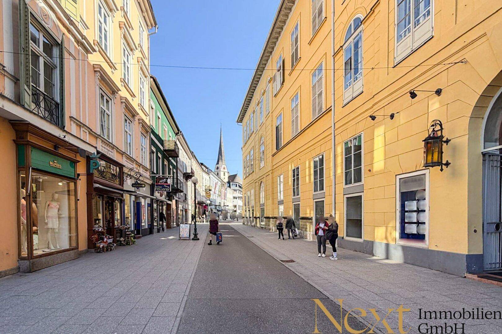 Belebte Einkaufsstraße mit historischen Gebäuden, Fußgängern und Geschäften unter blauem Himmel.