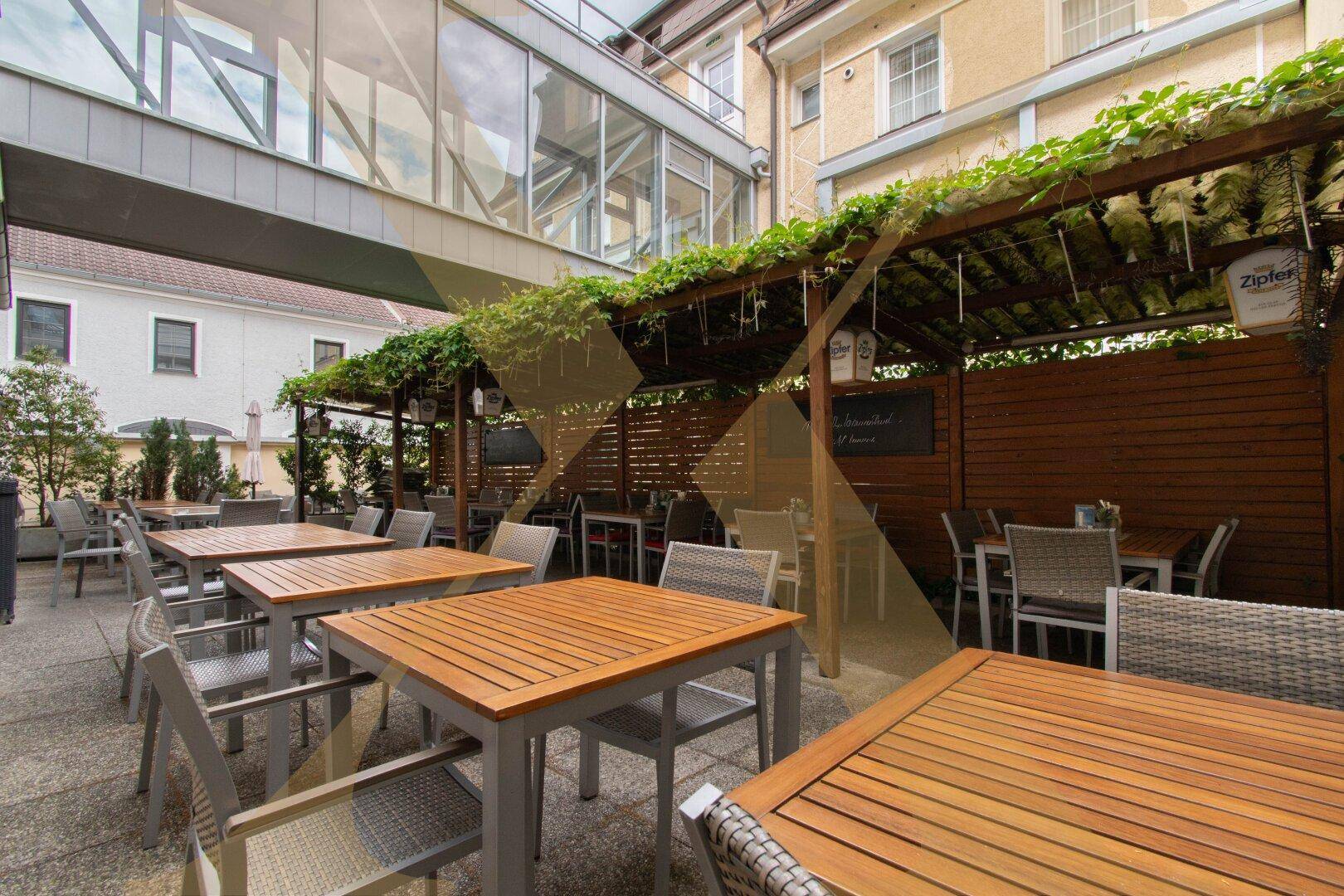 Außenbereich mit Holztischen und Stühlen unter einer begrünten Pergola.