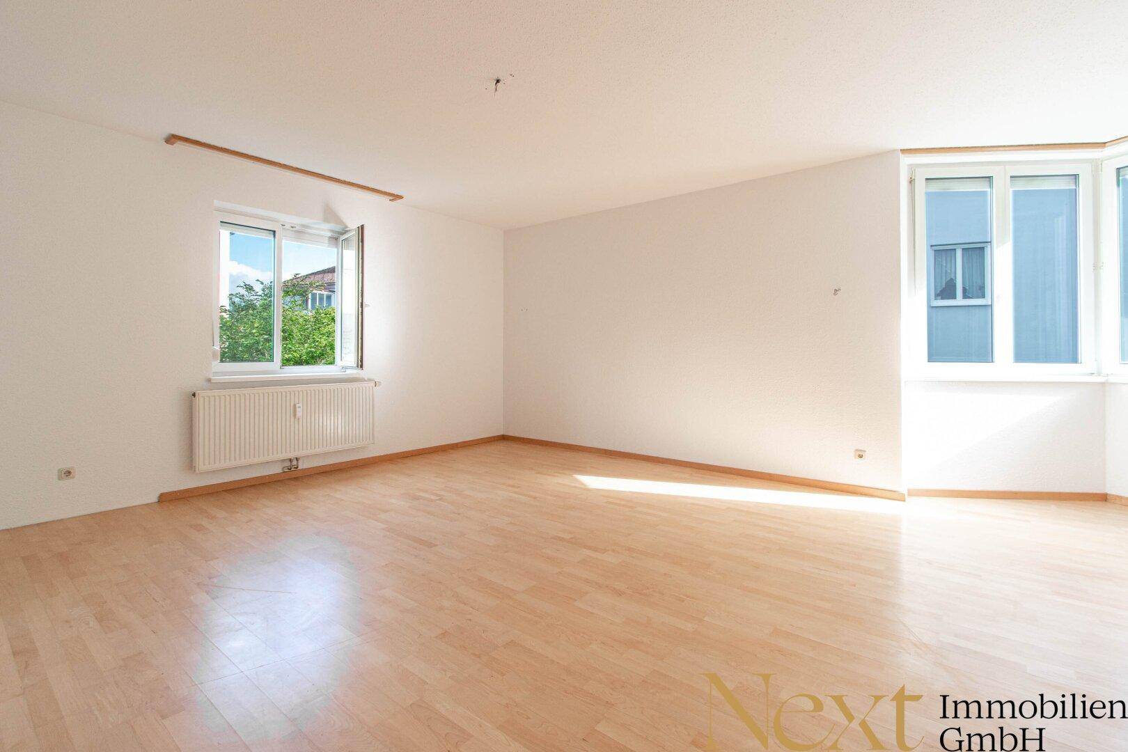 Großes, lichtdurchflutetes Zimmer mit Holzfußboden und mehreren Fenstern.