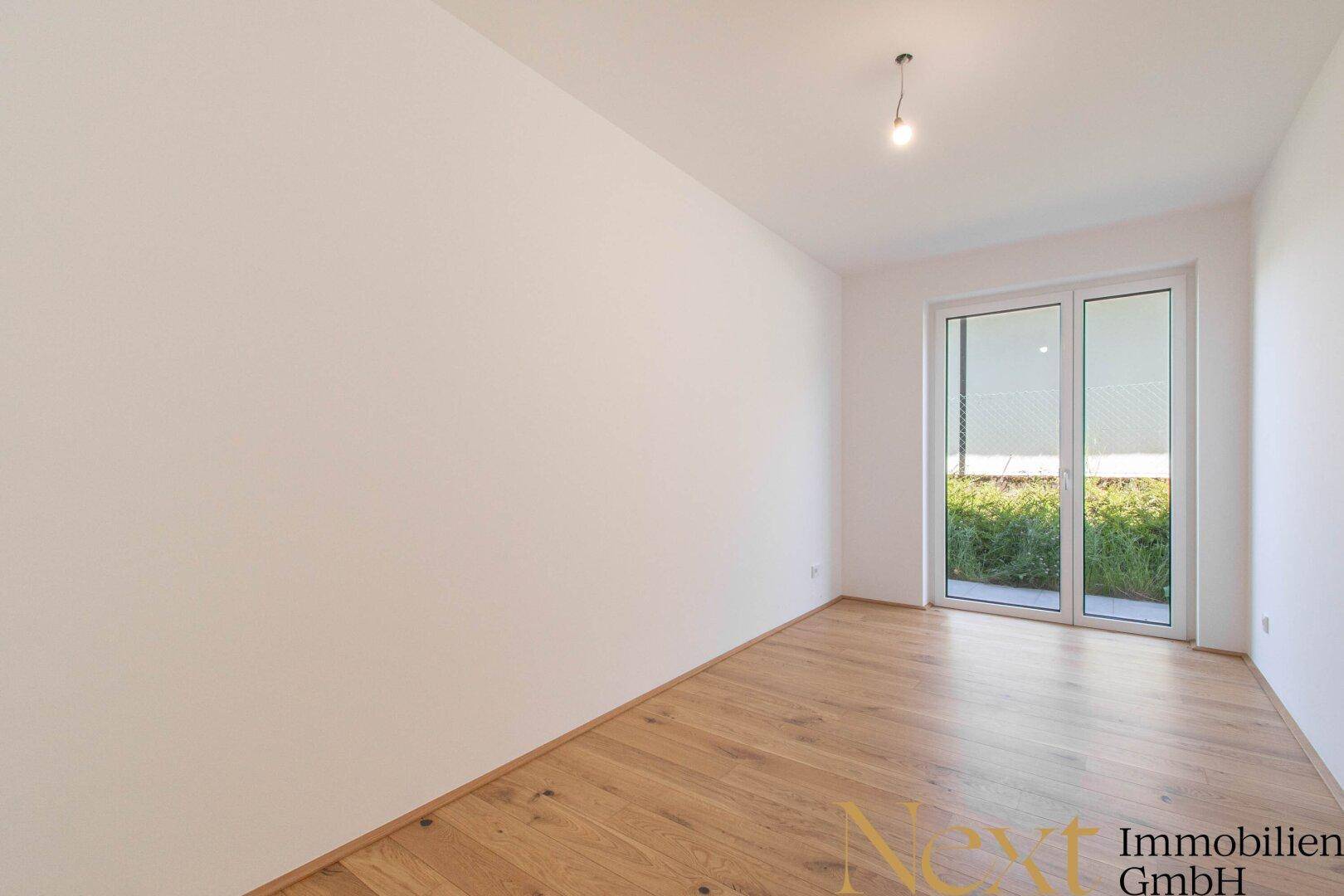 Leeres Zimmer mit Holzboden und großen Fenstern, die viel Licht hereinlassen und eine grüne Aussicht bieten.