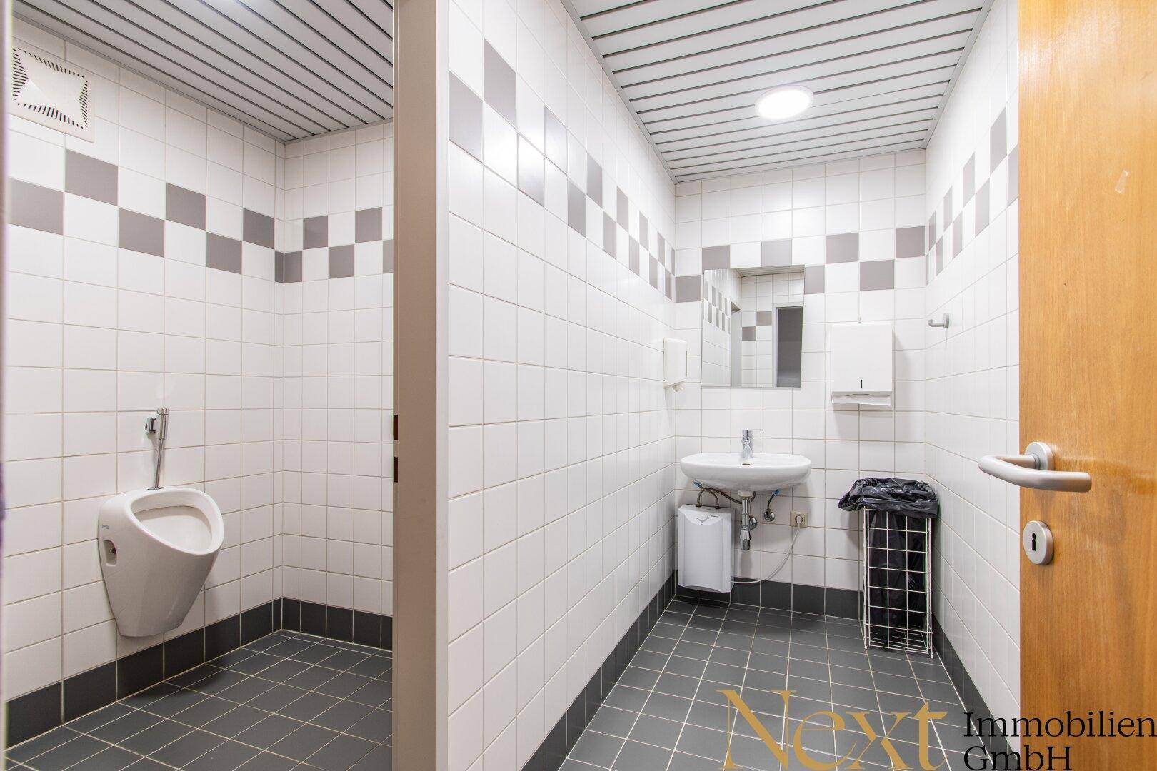 Herrenwaschraum mit Urinal, Waschbecken und weiß-grauen Fliesen an Wänden und Boden.