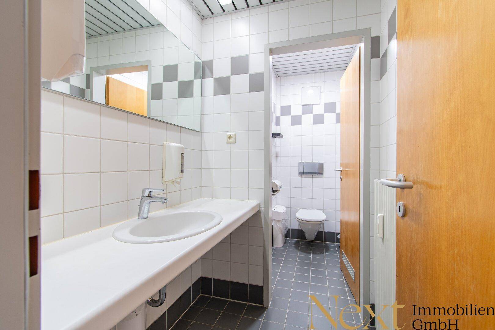 Waschraum mit langem Waschtisch, Spiegel und Zugang zu einer Toilettenkabine mit WC.