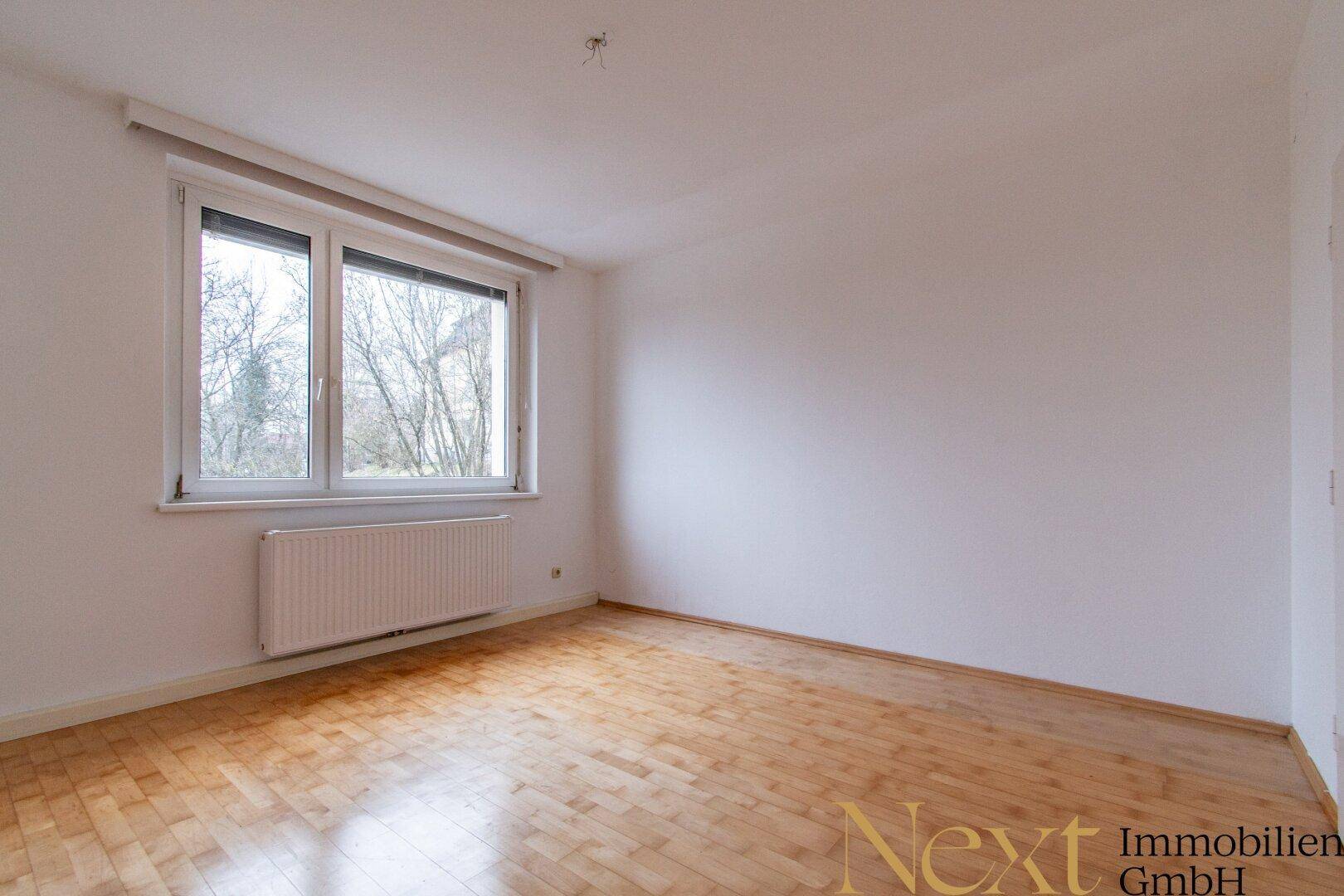 Leeres Zimmer mit Holzboden, großem Fenster und Heizkörper, bietet viel Tageslicht und Blick ins Grüne.