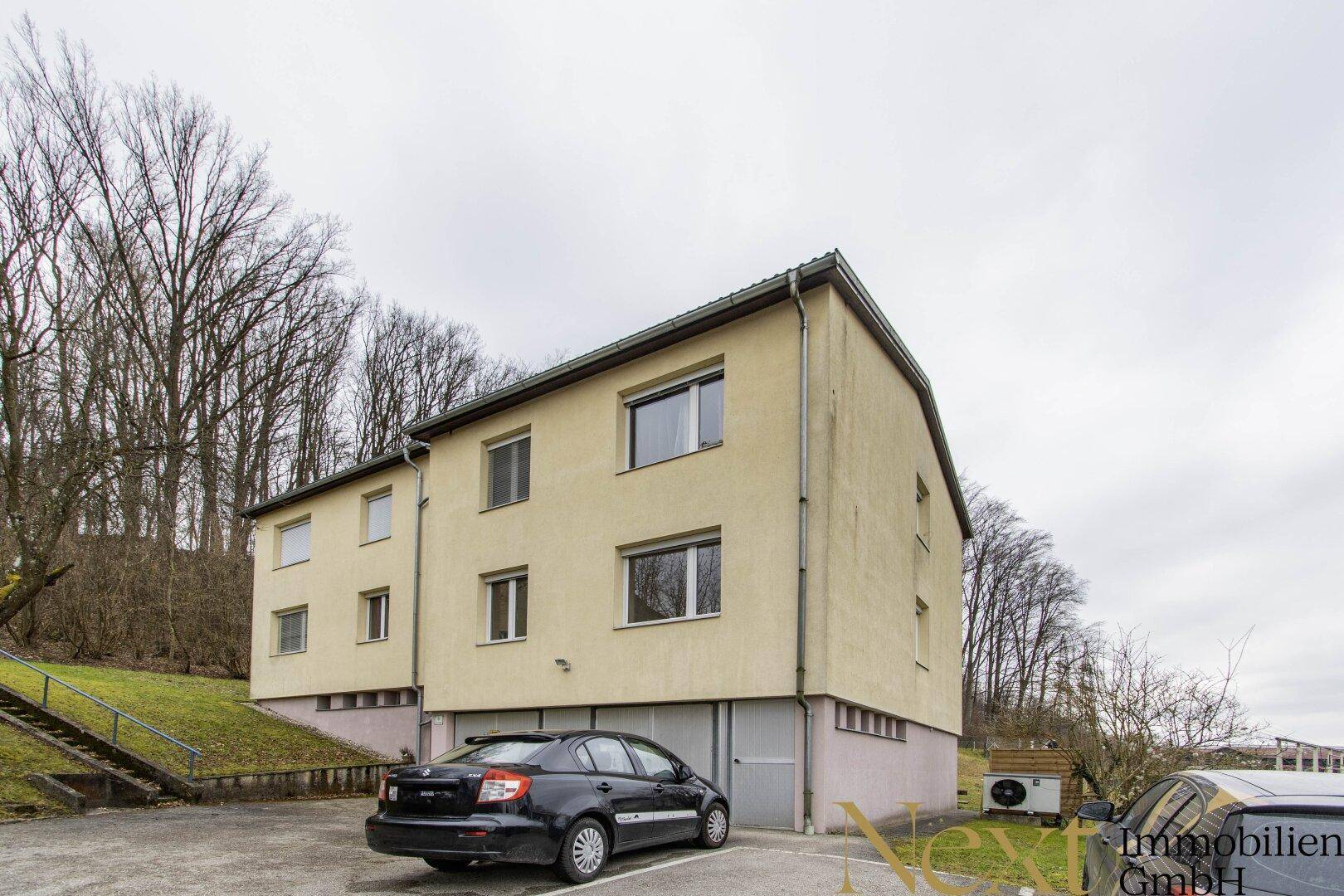Mehrstöckiges Gebäude mit gelber Fassade, Garage und Parkplätzen vor einem bewaldeten Hang.