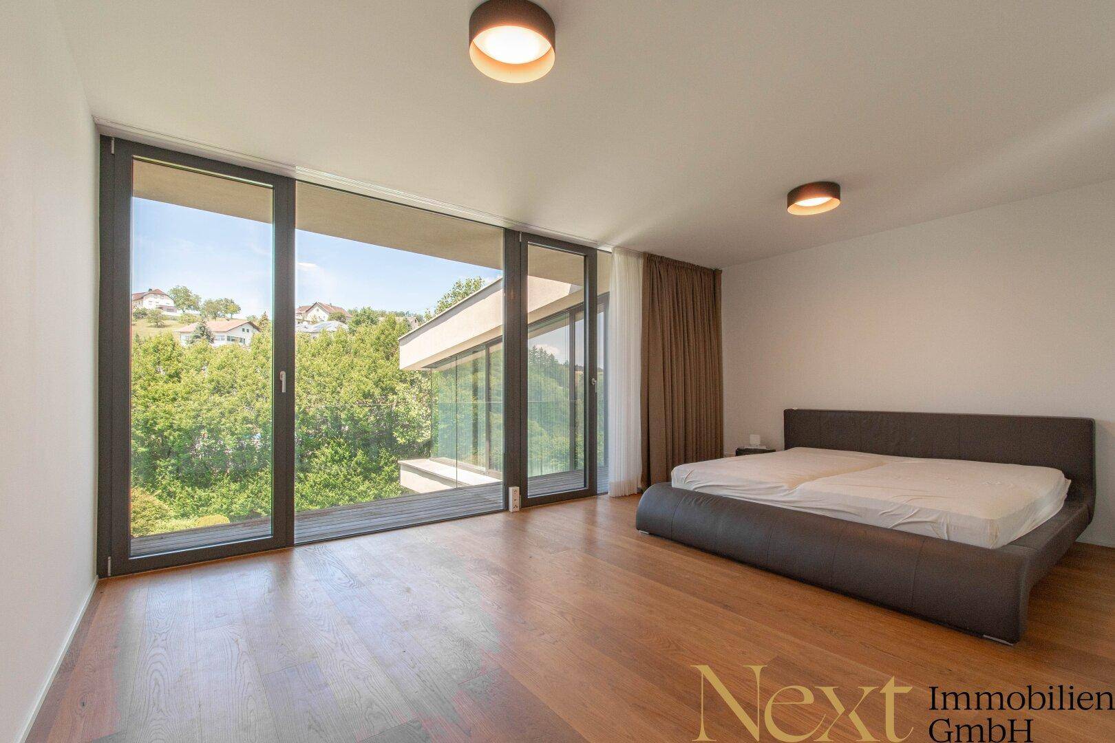 Schlafzimmer mit Holzboden und raumhohen Fenstern, die einen weiten Blick ins Grüne ermöglichen.