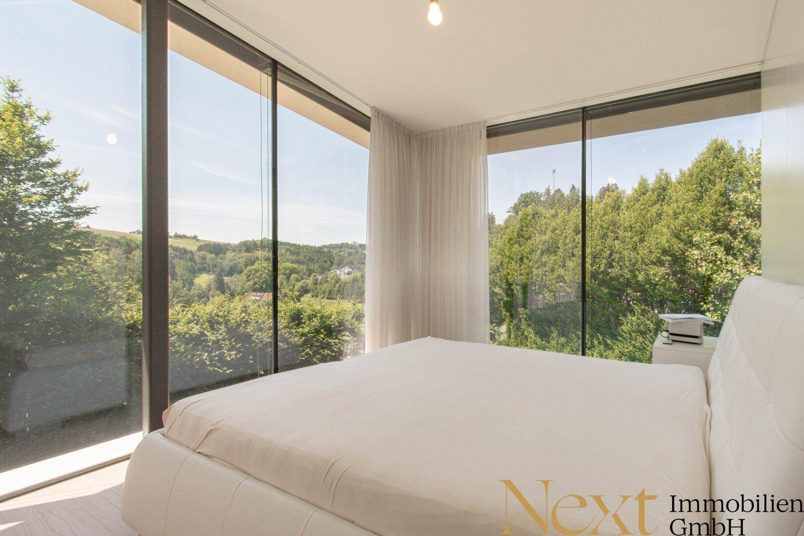 Helles Schlafzimmer mit weißem Bett und raumhohen Fenstern, die einen idyllischen Blick ins Grüne bieten.