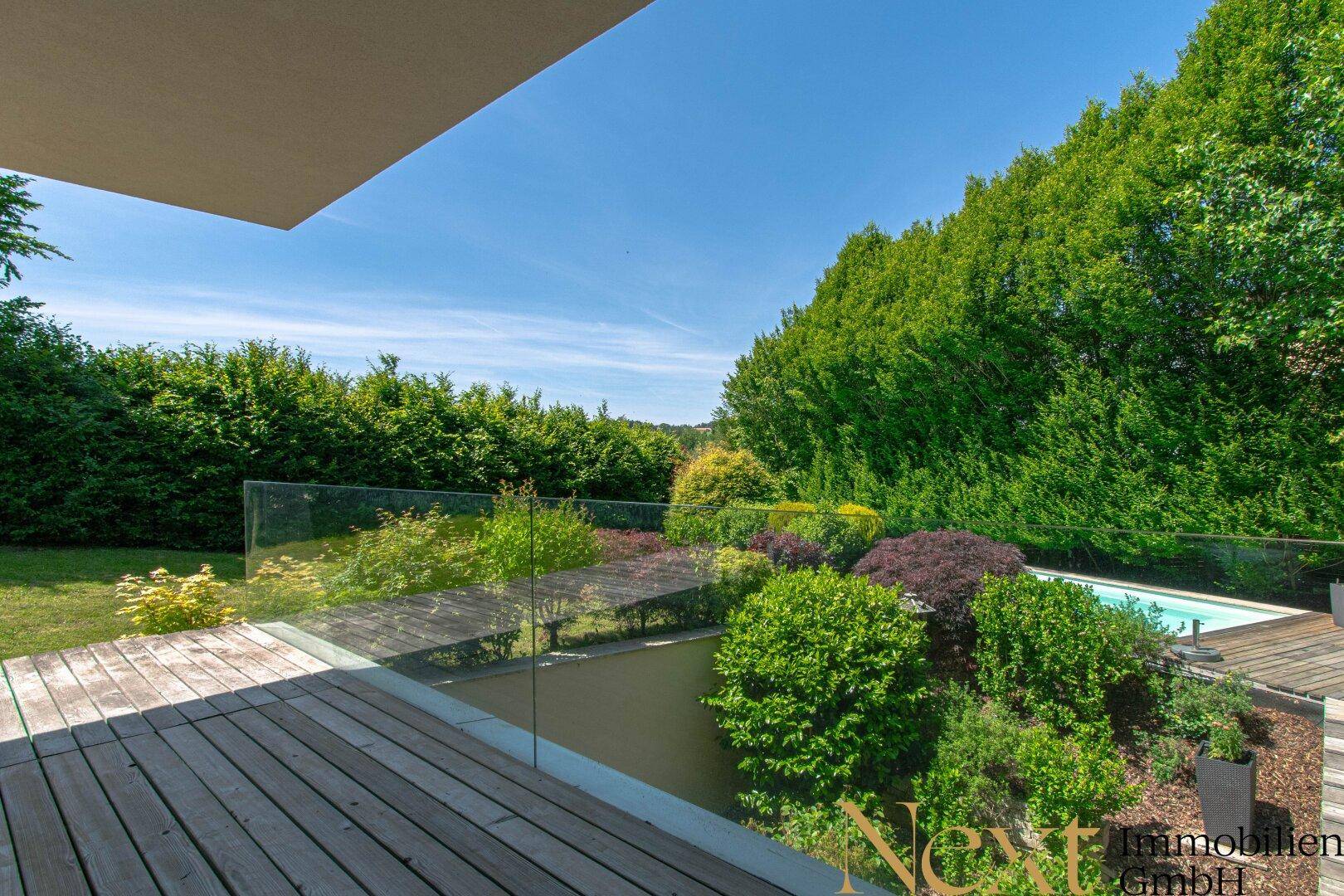 Geräumige Terrasse mit Glasgeländer und Holzdeck, mit Blick auf den Garten und den Poolbereich.