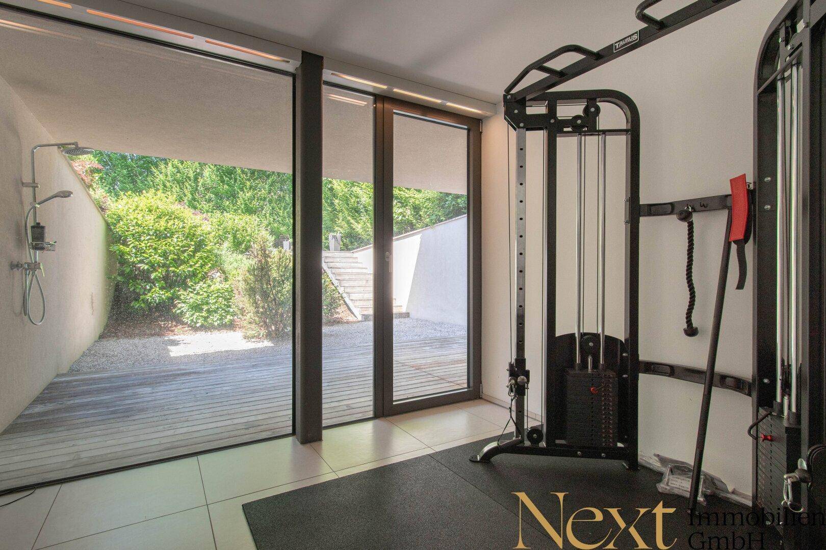 Moderner Fitnessraum mit Trainingsgeräten, großen Glasfronten und Zugang zu einem Außenbereich mit Dusche.