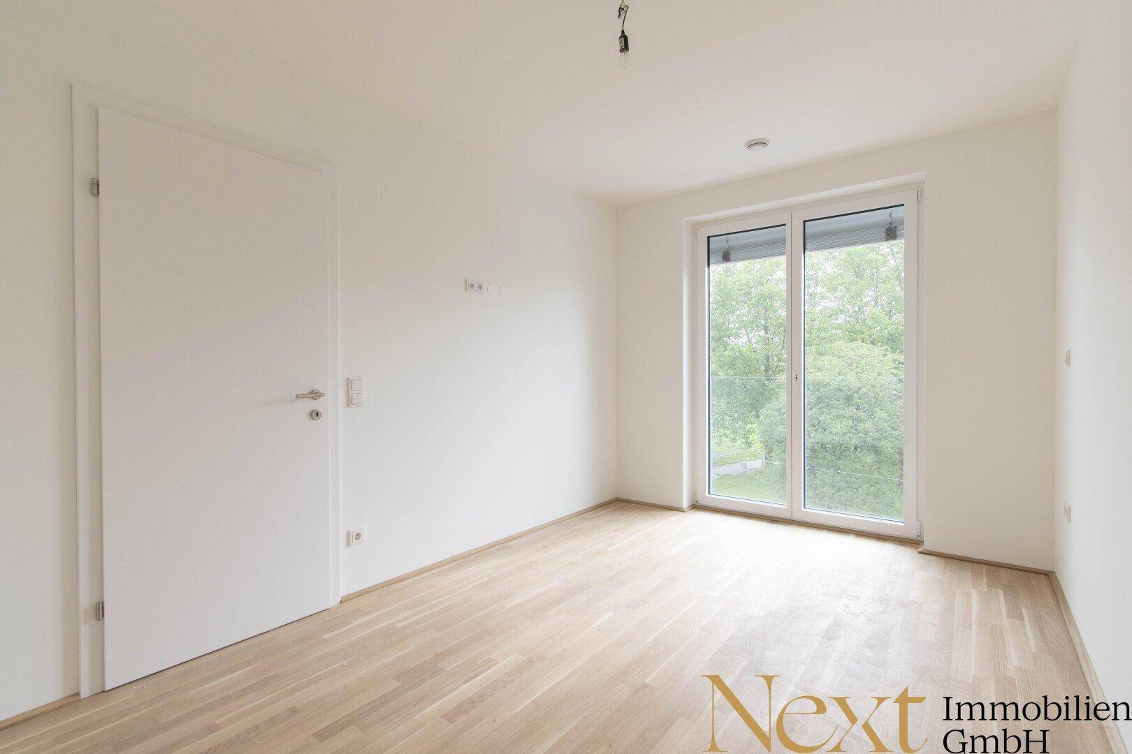 Helles Schlafzimmer mit Parkettboden und großem Fenster, das viel Tageslicht hereinlässt.
