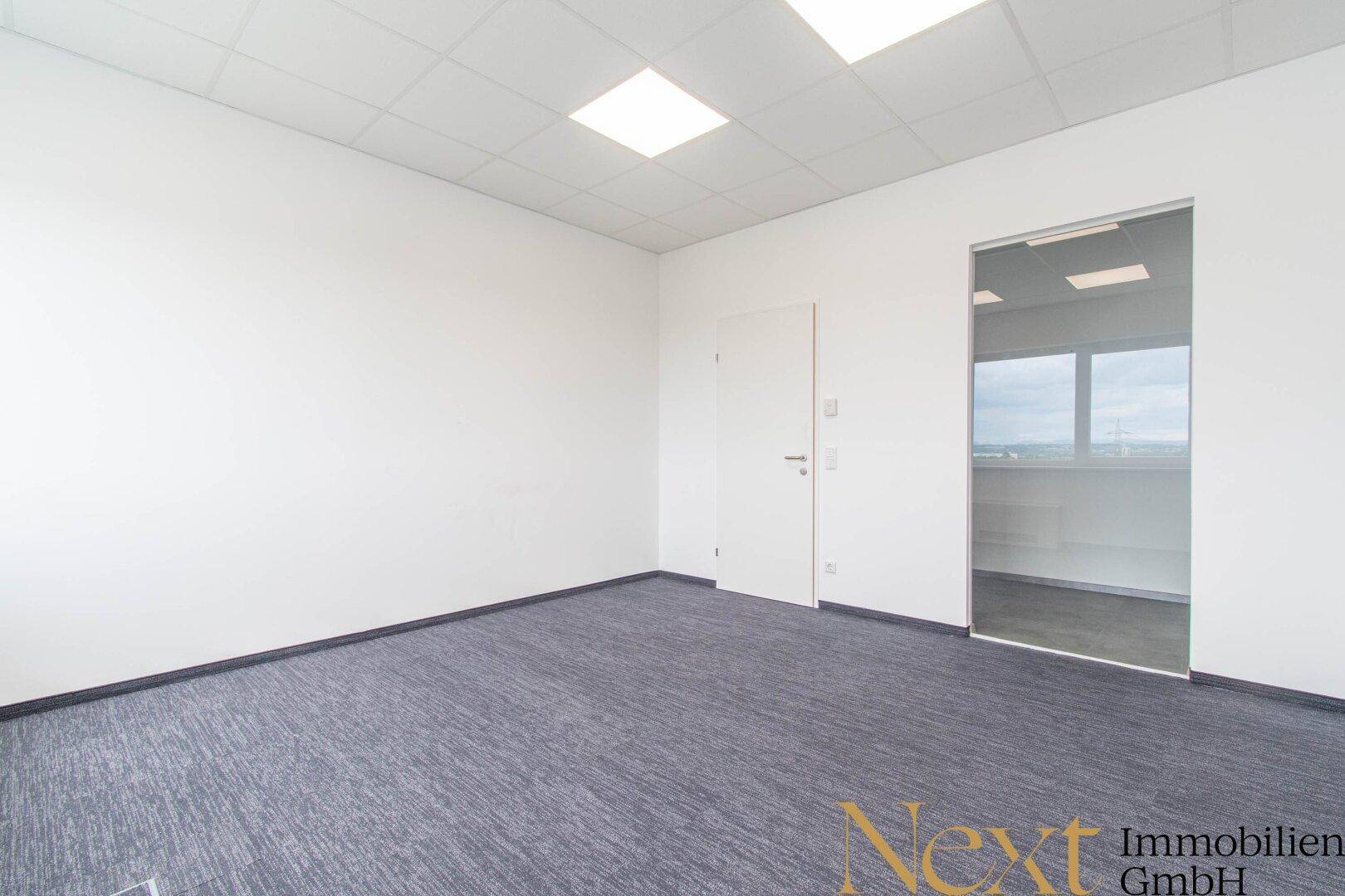 Leeres Büro mit Teppichboden und einer Glastür, die zu einem weiteren hellen Raum führt.