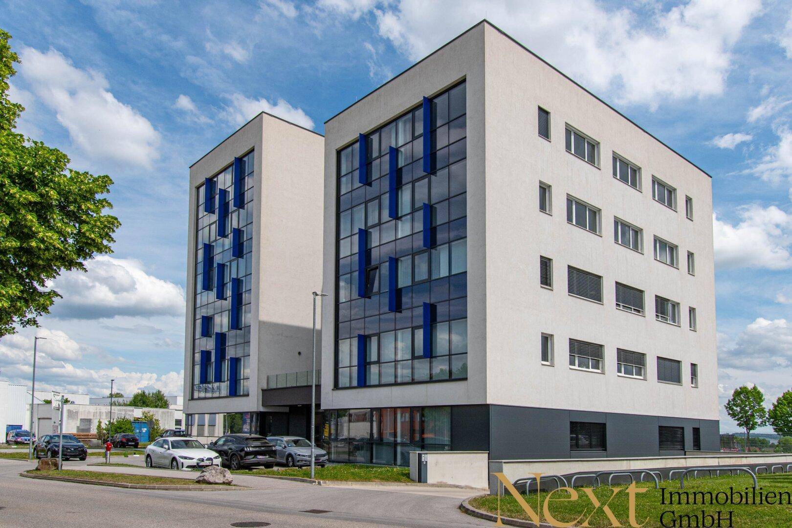 Modernes Bürogebäude mit weißer Fassade, großen Fenstern und blauen Akzenten unter blauem Himmel.