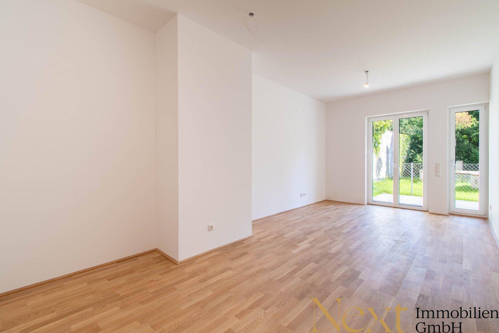 Heller, leerer Wohnraum mit Holzboden und großen Fenstern zum Gartenbereich.