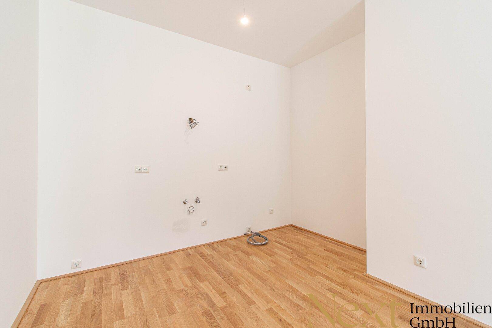 Unmöblierte Küche mit vorbereiteten Anschlüssen und Parkettboden in einem Neubau.