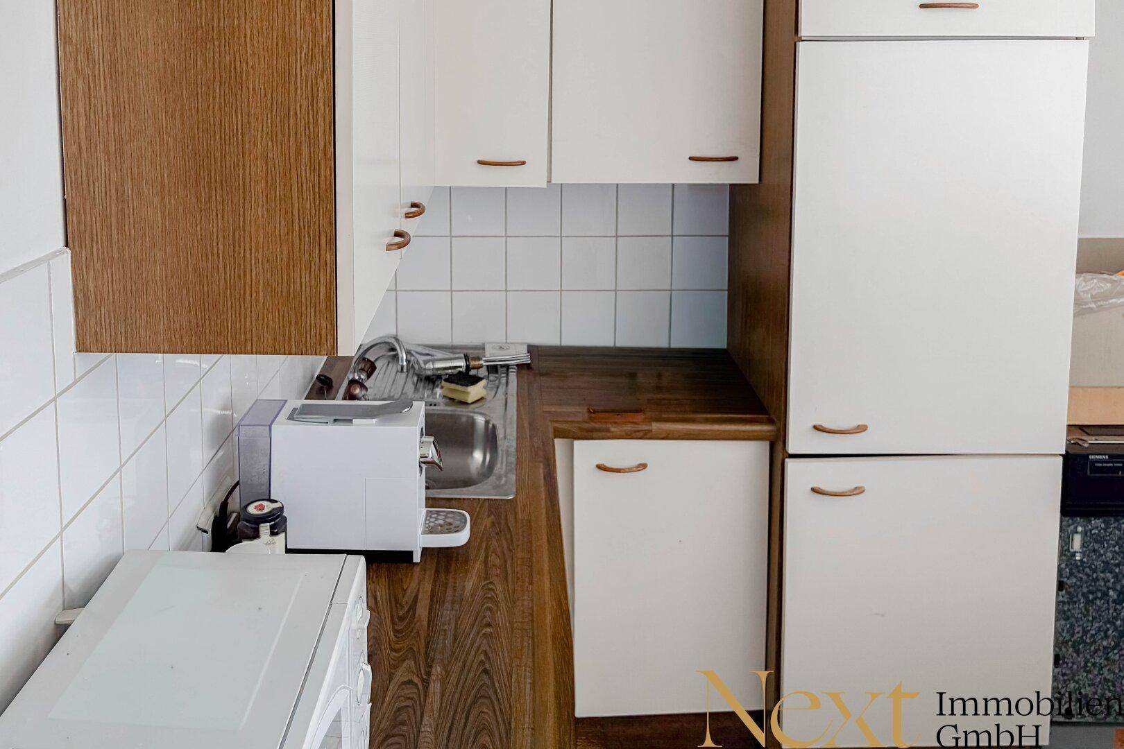Kleine Küchenzeile mit weißen Schränken, Holzoptik-Arbeitsplatte und integriertem Kühlschrank, funktional eingerichtet.