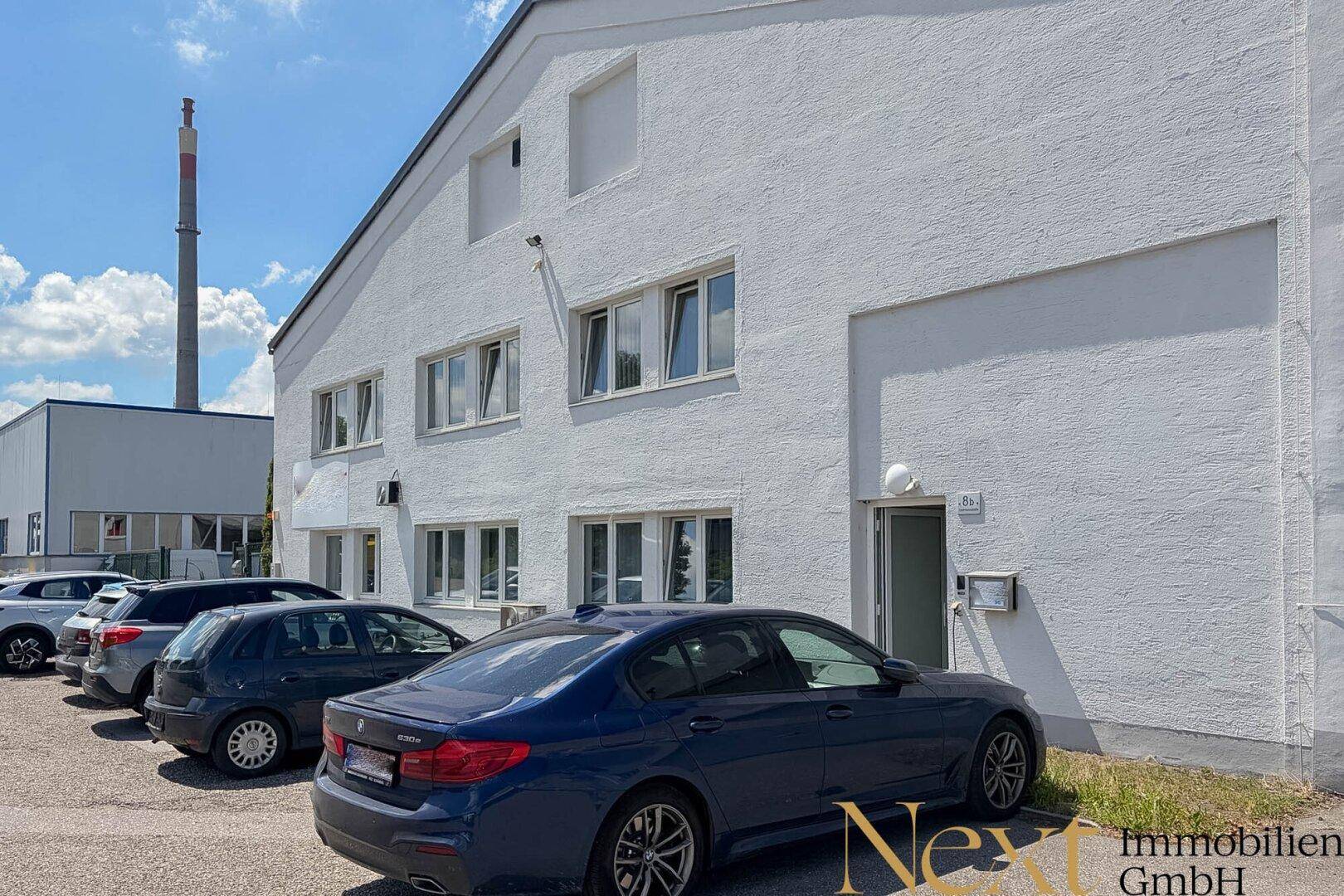 Seitenansicht des weißen Gewerbegebäudes mit Parkplätzen und einem hohen Schornstein im Hintergrund.