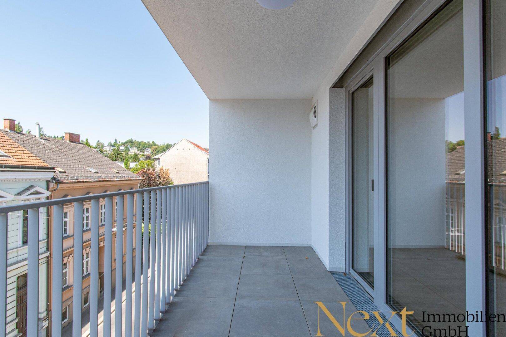 Geräumiger Balkon mit Fliesenboden und Schiebetür, bietet einen Blick auf die Straße und Nachbargebäude.