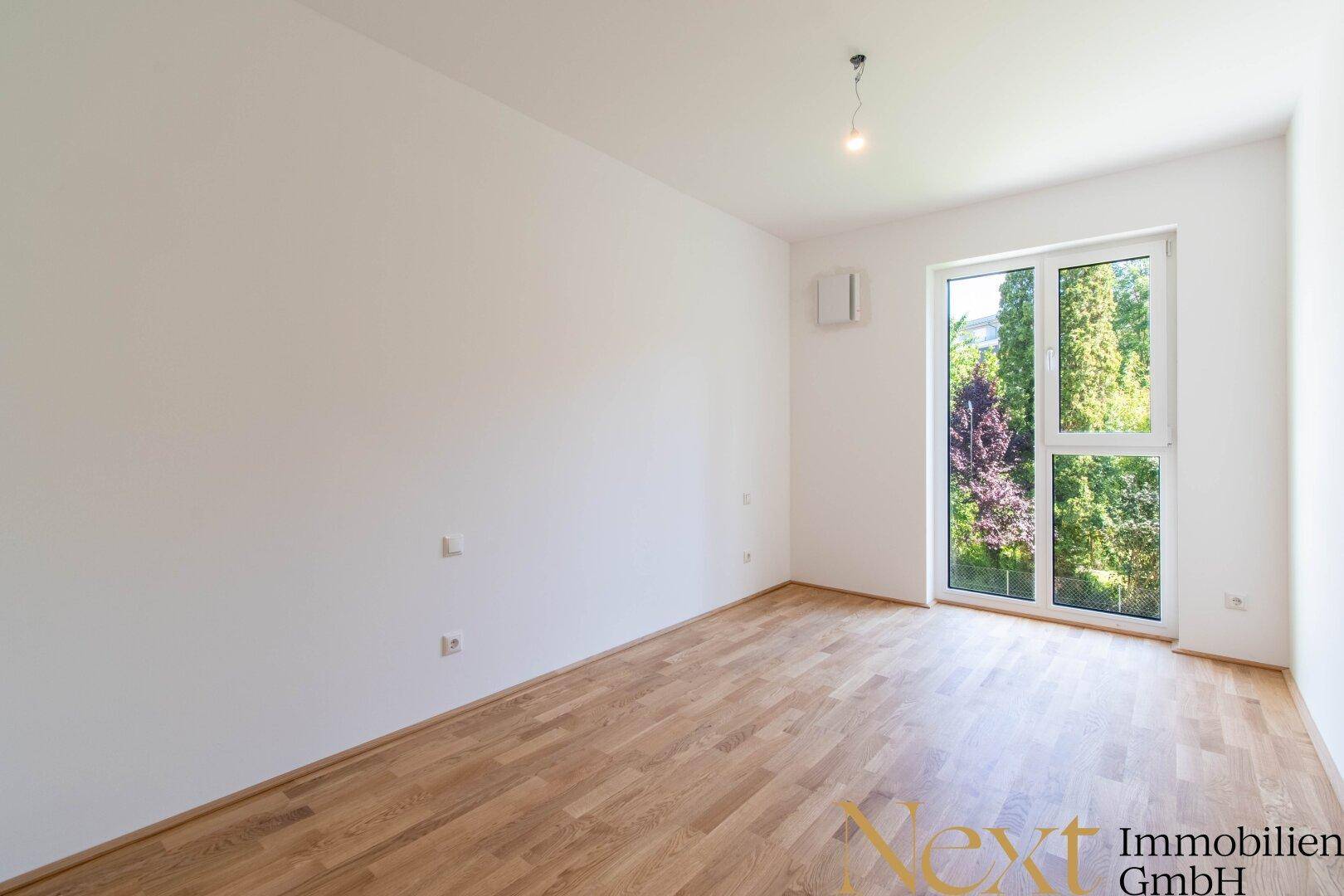 Unmöbliertes Zimmer mit hellem Parkettboden und großem Fenster, das viel natürliches Licht hereinlässt.