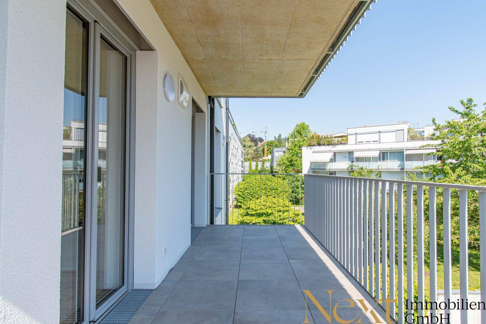 Heller Balkon mit Fliesenboden und Blick auf die grüne Umgebung und weitere Wohngebäude.