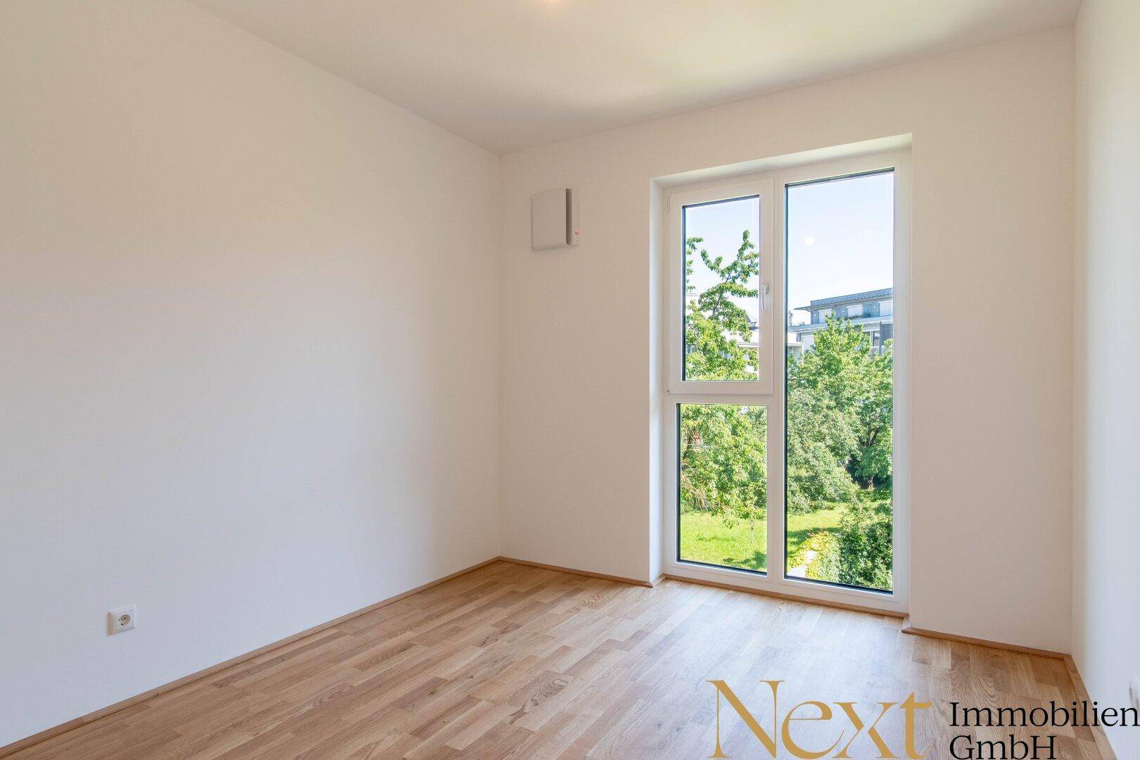 Helles Schlafzimmer mit Parkettboden und einem großen Fenster mit Blick ins Grüne.