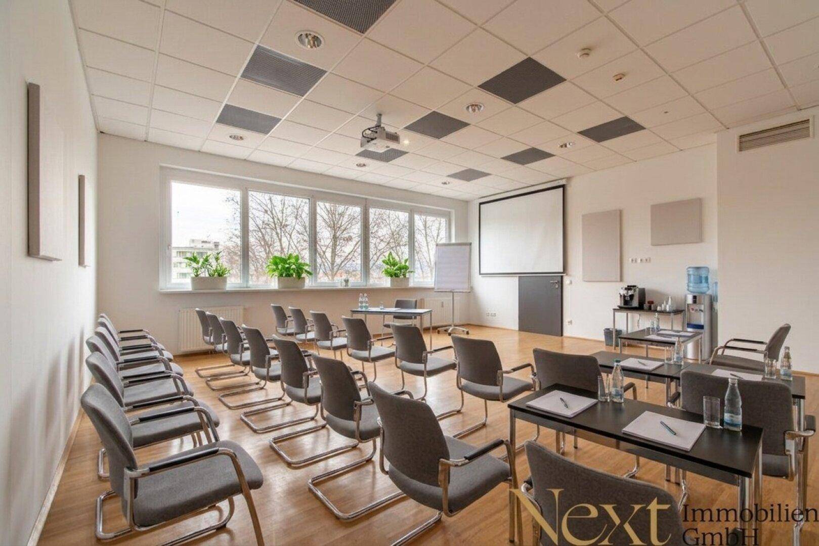 Großer Konferenzraum mit vielen Stühlen, Tischen, einem Beamer und großen Fenstern für Besprechungen.