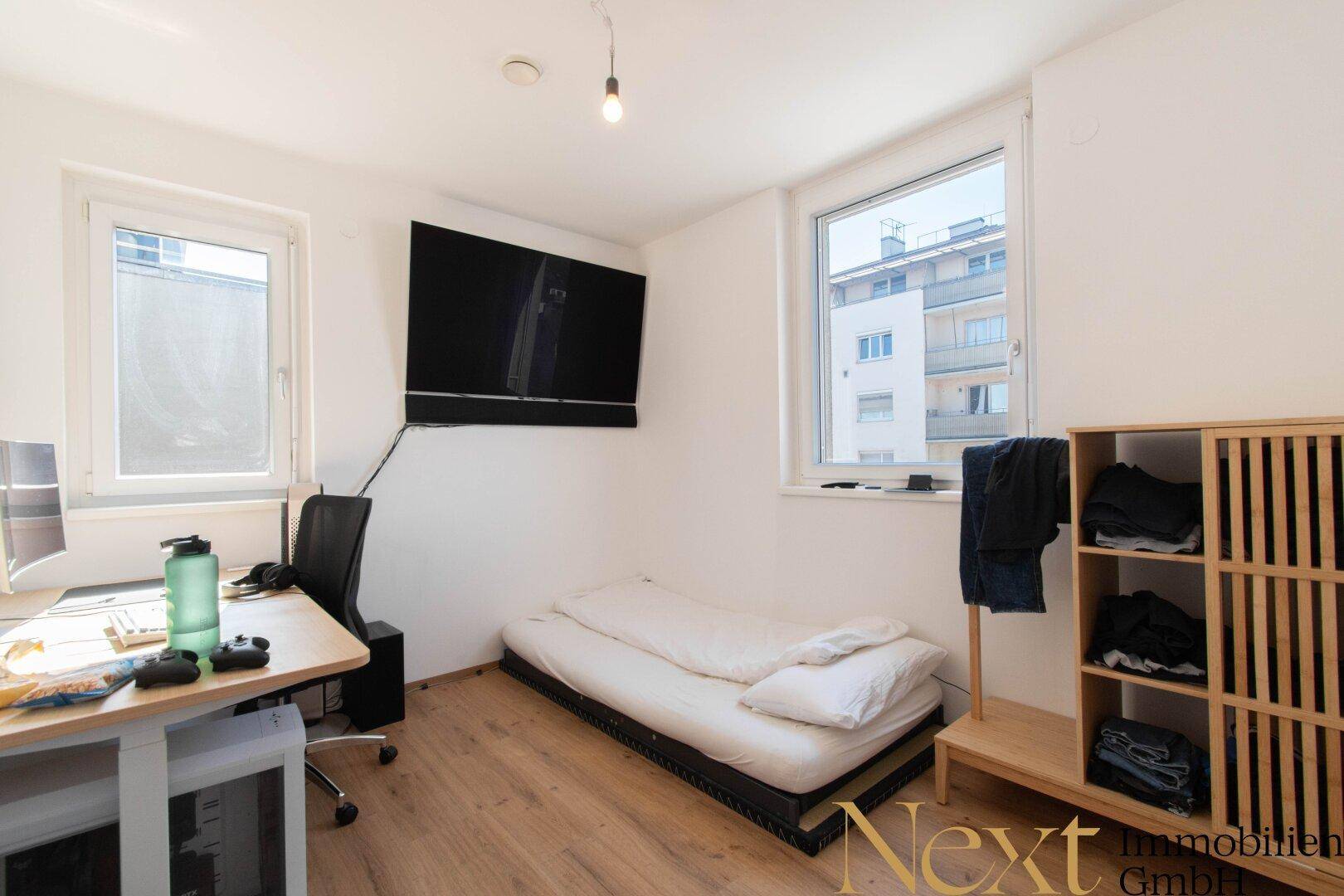 Helles Schlafzimmer mit Wandfernseher, Schreibtisch und praktischem Stauraumregal.