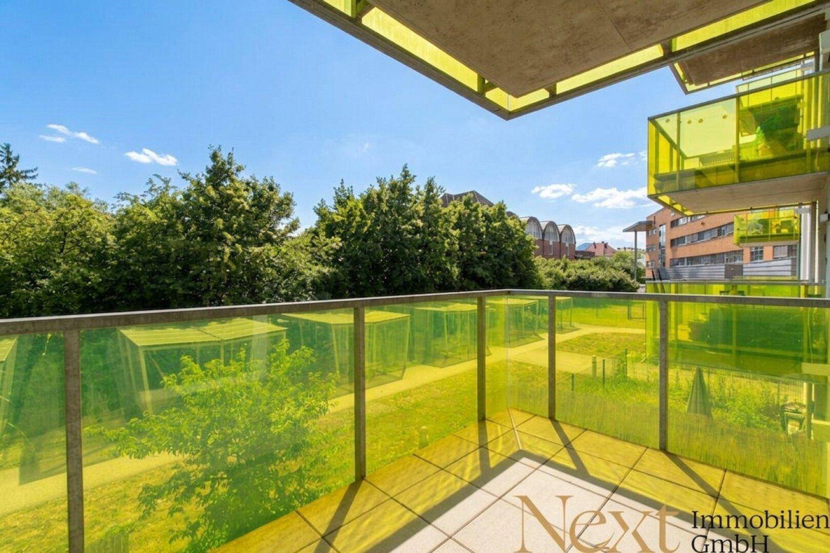 Heller Balkon mit gelb-grüner Glasbrüstung und Ausblick auf Bäume und umliegende Gebäude.