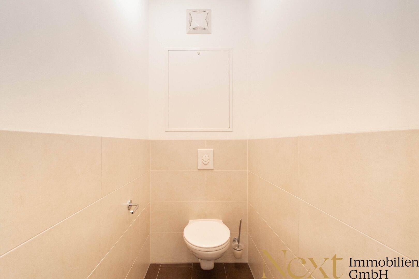 Separates WC mit beigefarbenen Fliesen, Wand-WC und Abluftsystem.