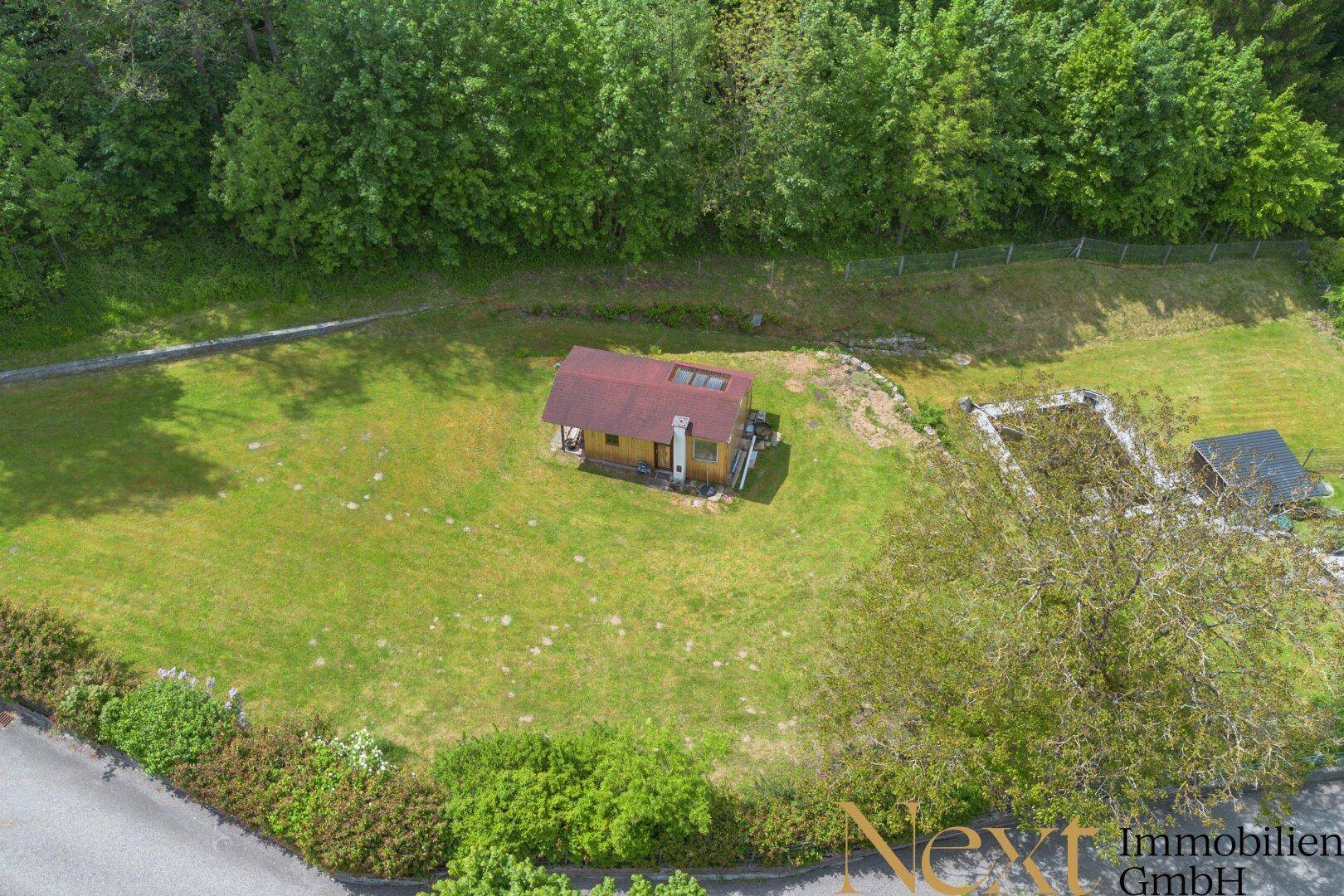 Luftaufnahme des Anwesens mit einem kleinen Holzhaus, weitläufiger Grünfläche und umgebenden Bäumen.