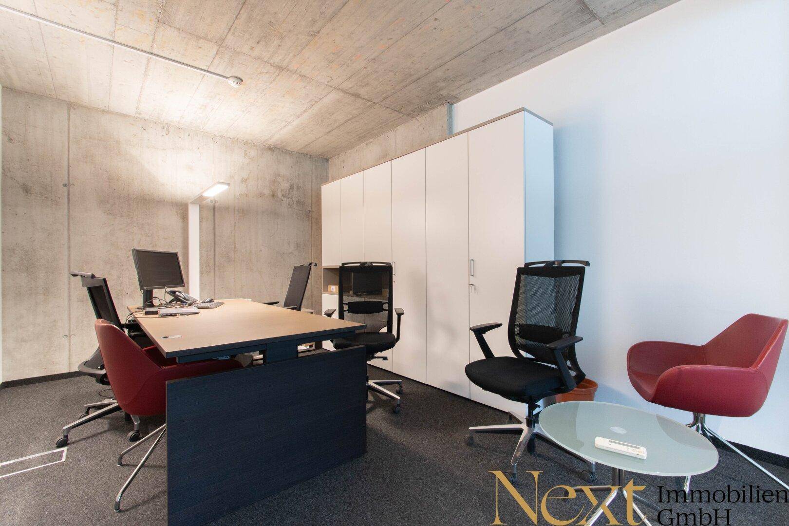 Helles Büro mit Schreibtisch, ergonomischen Stühlen, weißen Aktenschränken und einem roten Sessel für Besucher.