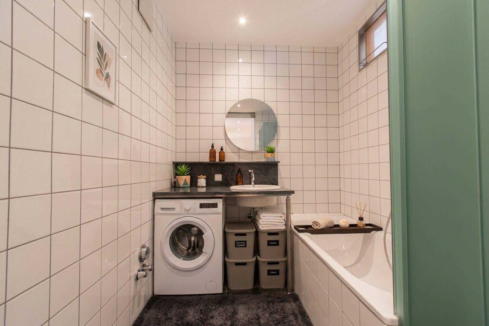 Helles Badezimmer mit weißen Fliesen, Badewanne, Waschbecken und integrierter Waschmaschine.