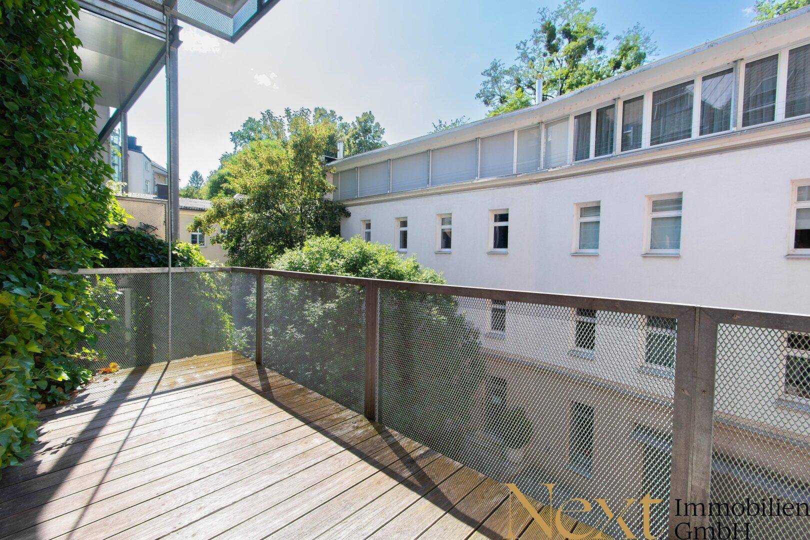 Heller Balkon mit Holzfußboden, Metallgeländer und Blick auf grüne Bäume und ein Gebäude.