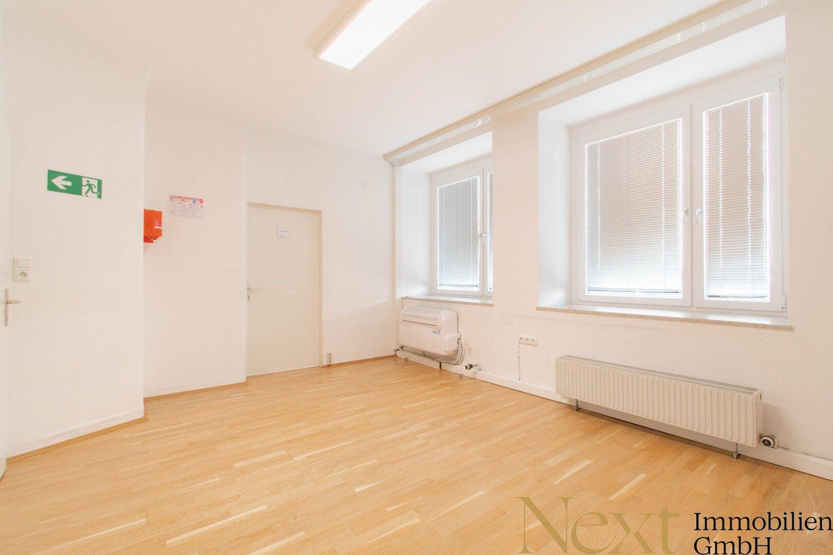 Geräumiger Büroraum mit Holzfußboden, mehreren Fenstern und Notausgangsschild.