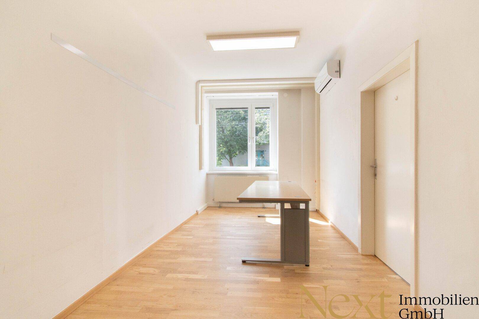 Schmaler Büroraum mit einem Schreibtisch, Klimaanlage und Fenster mit Blick ins Grüne.