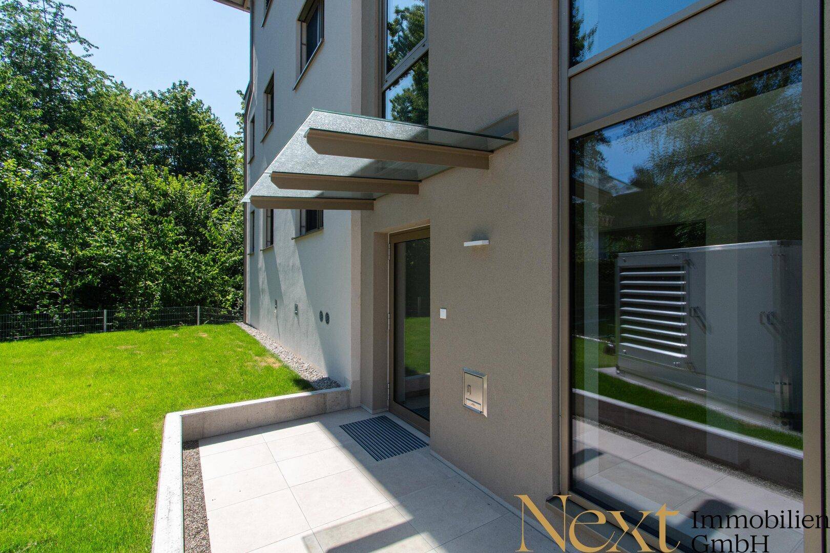 Eingangsbereich des Gebäudes mit kleiner Terrasse, Rasenfläche und moderner Wärmepumpe.