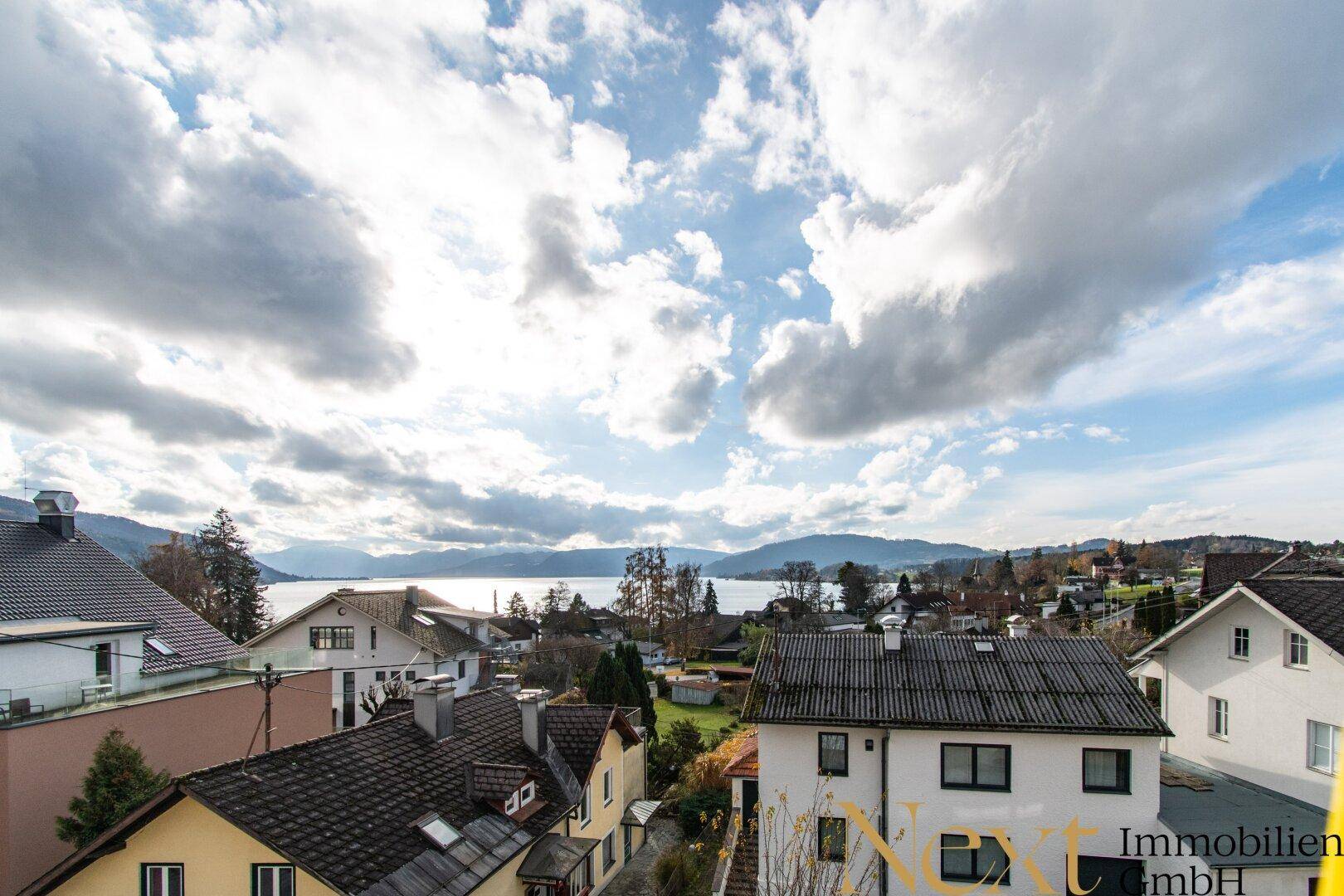 Panoramablick über eine Siedlung mit See und Bergen unter einem bewölkten Himmel.