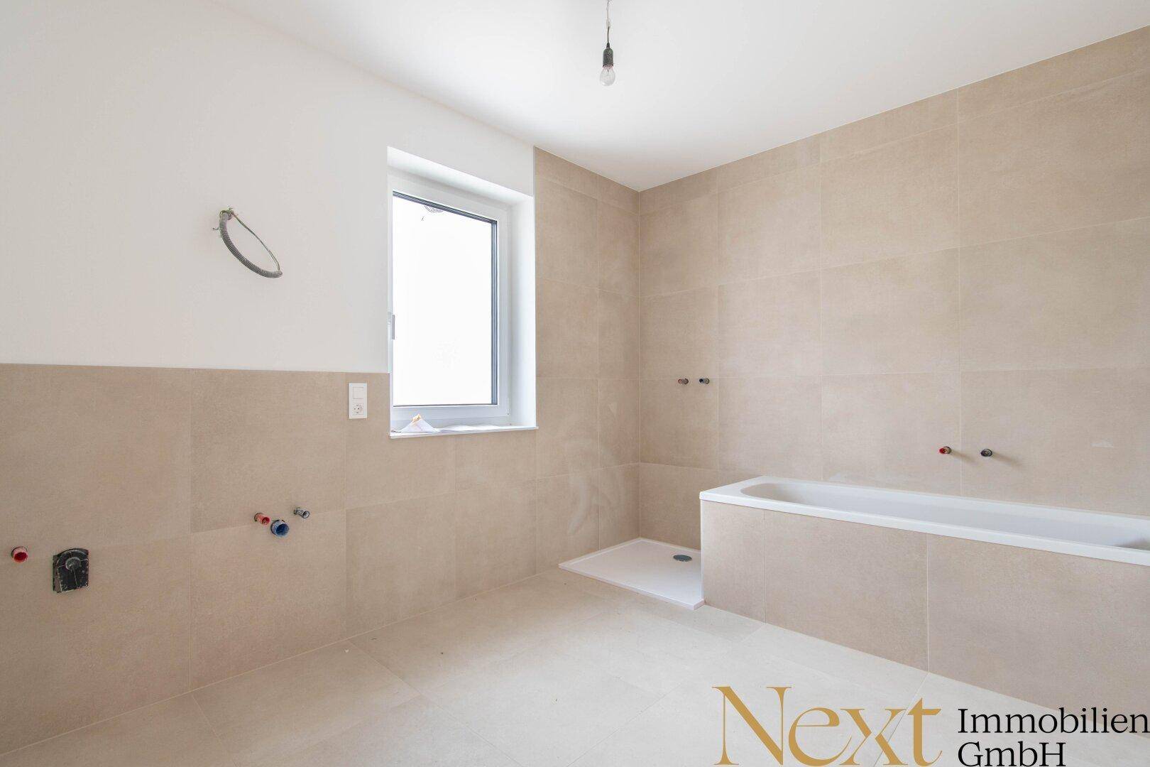 Helles Badezimmer mit beigefarbenen Fliesen, Badewanne, Duschbereich und Fenster.