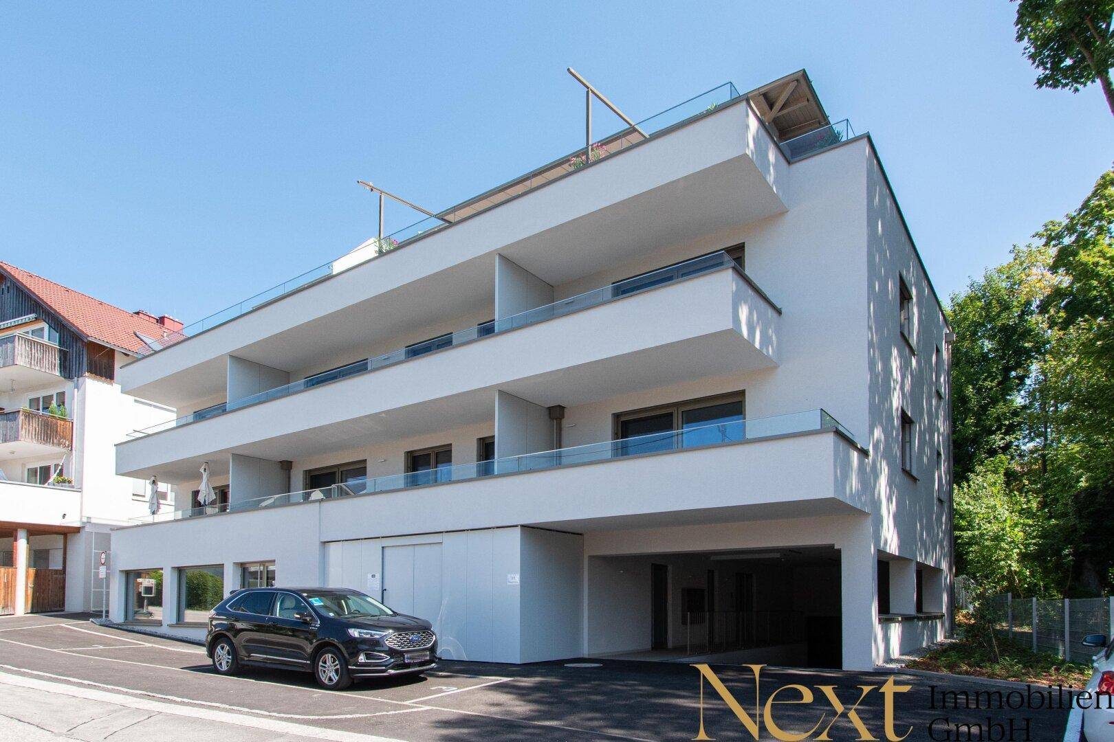 Modernes weißes Mehrfamilienhaus mit Balkonen und Tiefgaragenzufahrt, umgeben von Grünflächen.